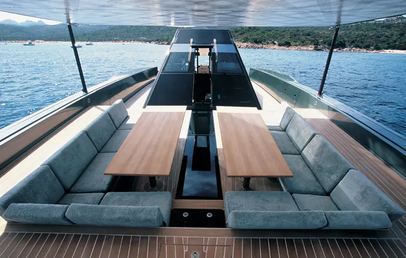Sunken seating area on the foredeck with two teak tables and grey cushions, the sea and a wooded coast ahead