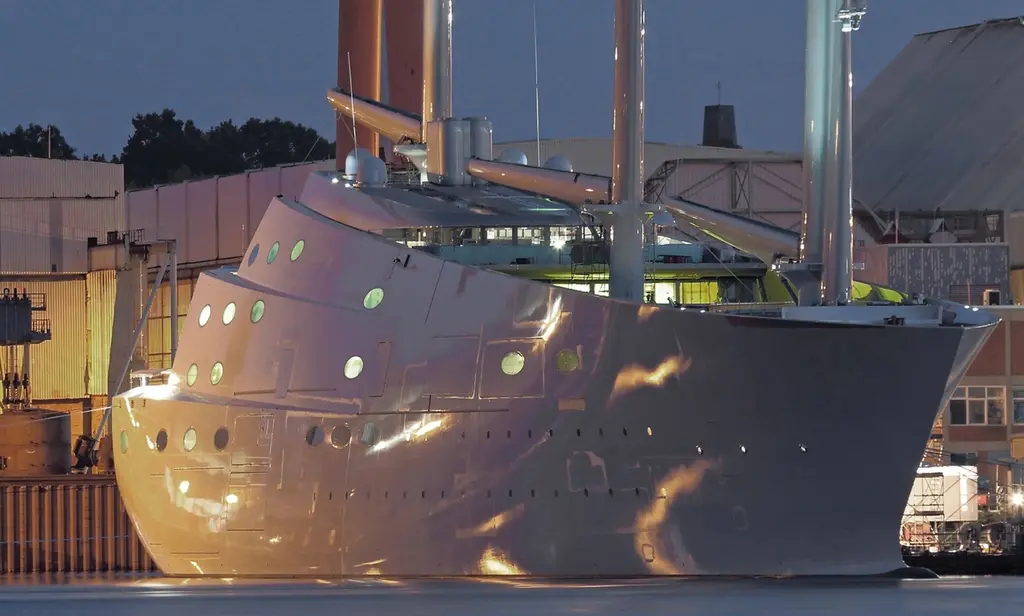 The high bow and oval windows of Sailing Yacht A alongside a shipyard quay at dusk