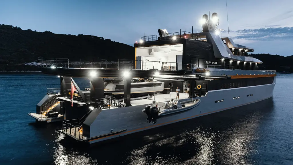 Nebula at anchor at dusk, seen from the starboard quarter: the lit helicopter hangar open on the upper deck, a white catamaran tender under the helideck and a RIB with outboard engines on the side deck