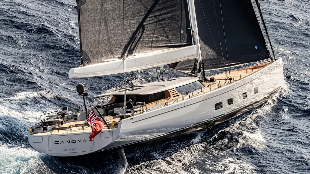 Canova seen from astern and above, sailing upwind, with the long hardtop over the cockpit and a red ensign at the stern