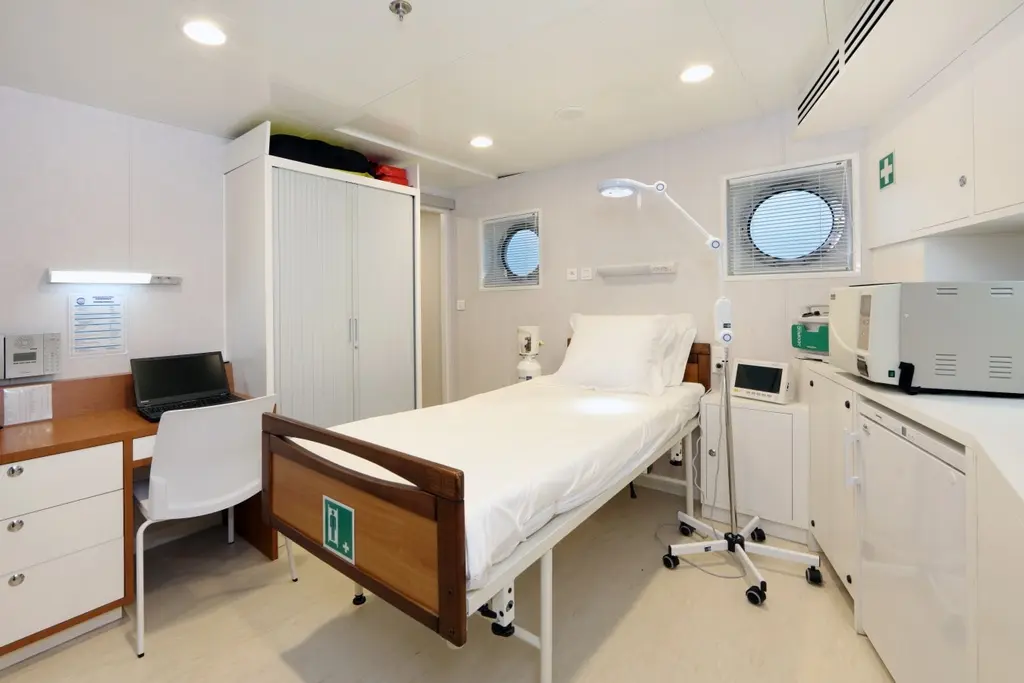 Ship's hospital with a medical bed, an examination lamp, a desk with a laptop and white cabinets