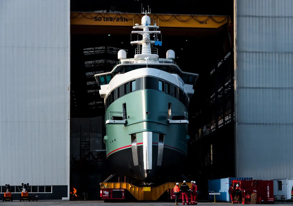 Anawa's bow emerging head-on from the doors of a build hall on a yellow transporter, with workers in red overalls in front