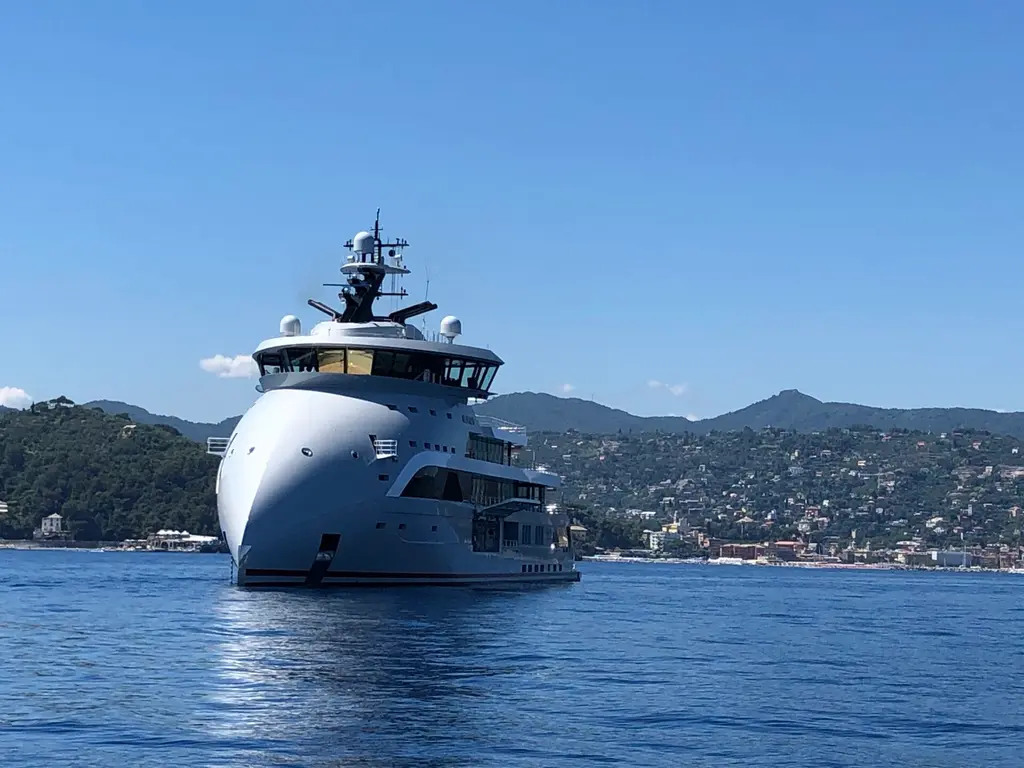 Olivia O at anchor from the port bow, three-quarter view, with a hillside town behind