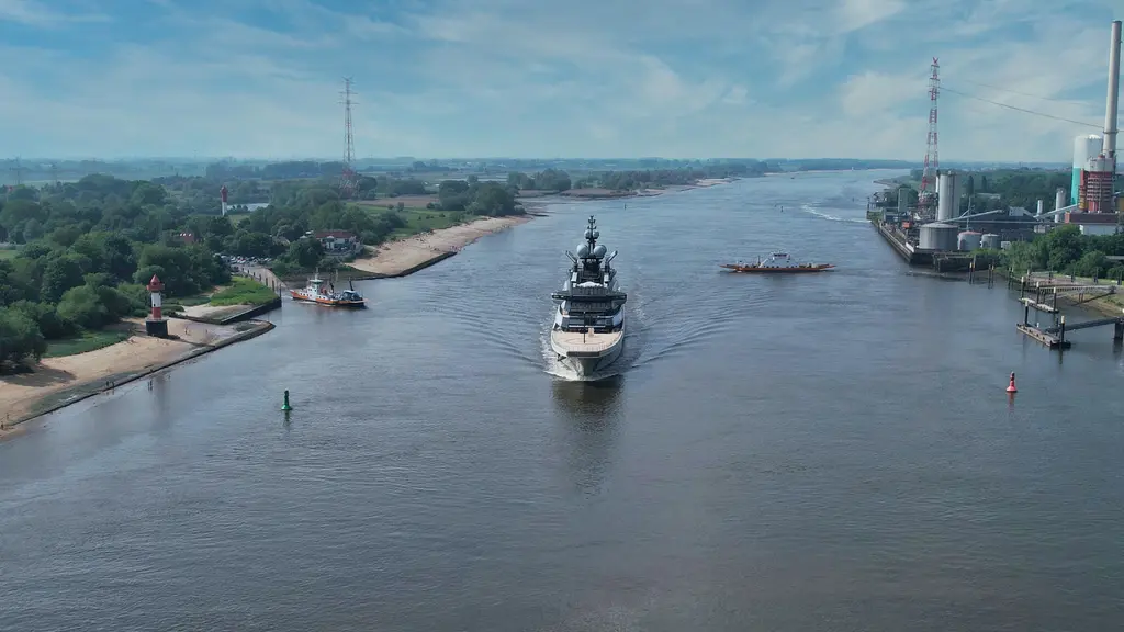 Aerial view of Nord heading towards the camera down a wide river, with a ferry and an industrial quay alongside