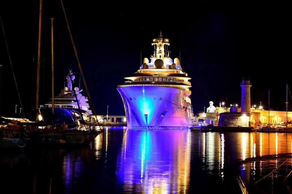 Dilbar's bow lit in blue and purple at night in a harbour beside a lighthouse