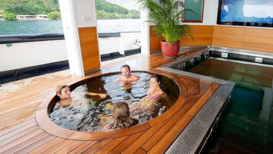 A round hot tub set into the teak deck with guests in it, next to a glass-sided pool
