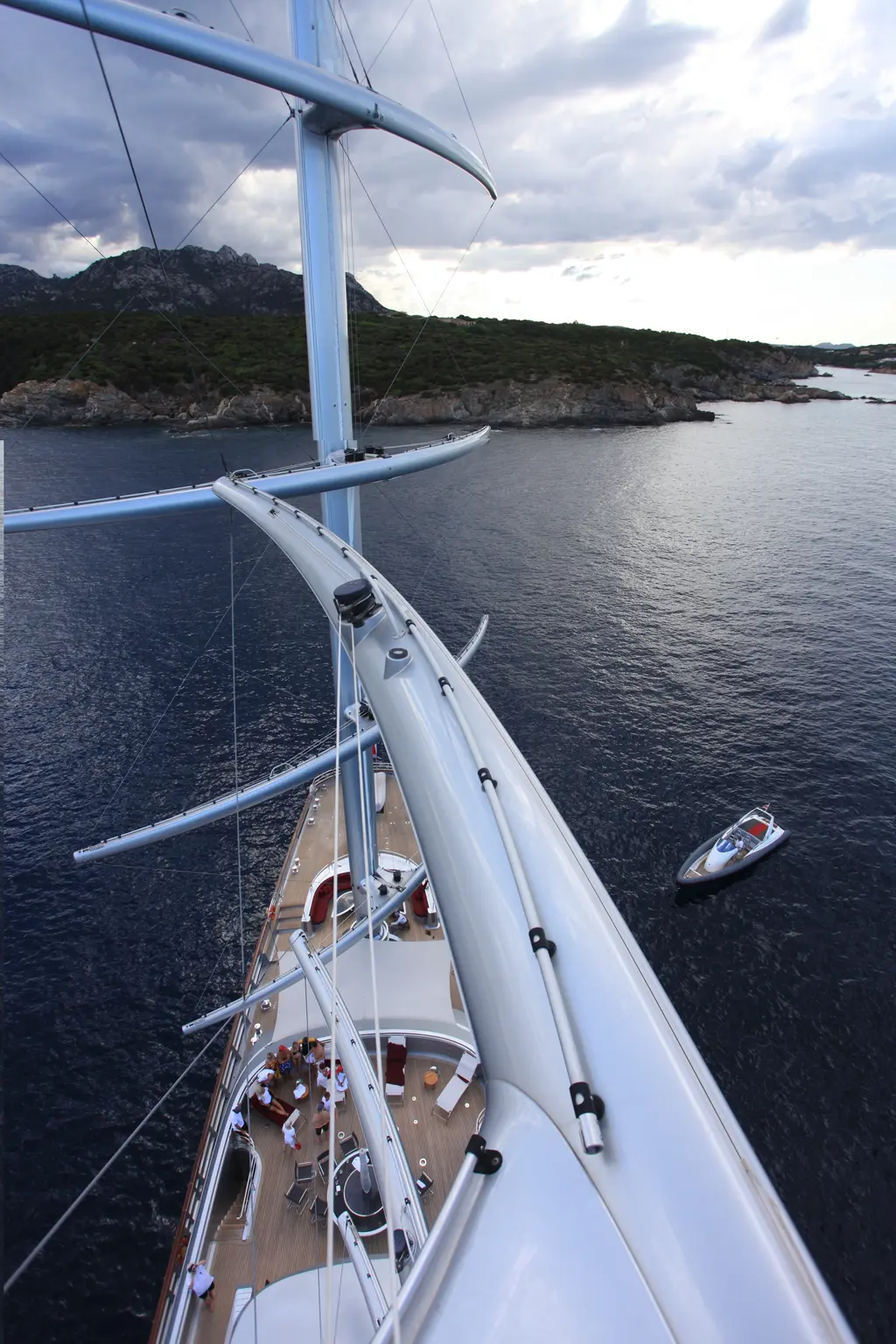 View down from high on a mast: curved yards with furled sails, the teak deck below and a tender alongside