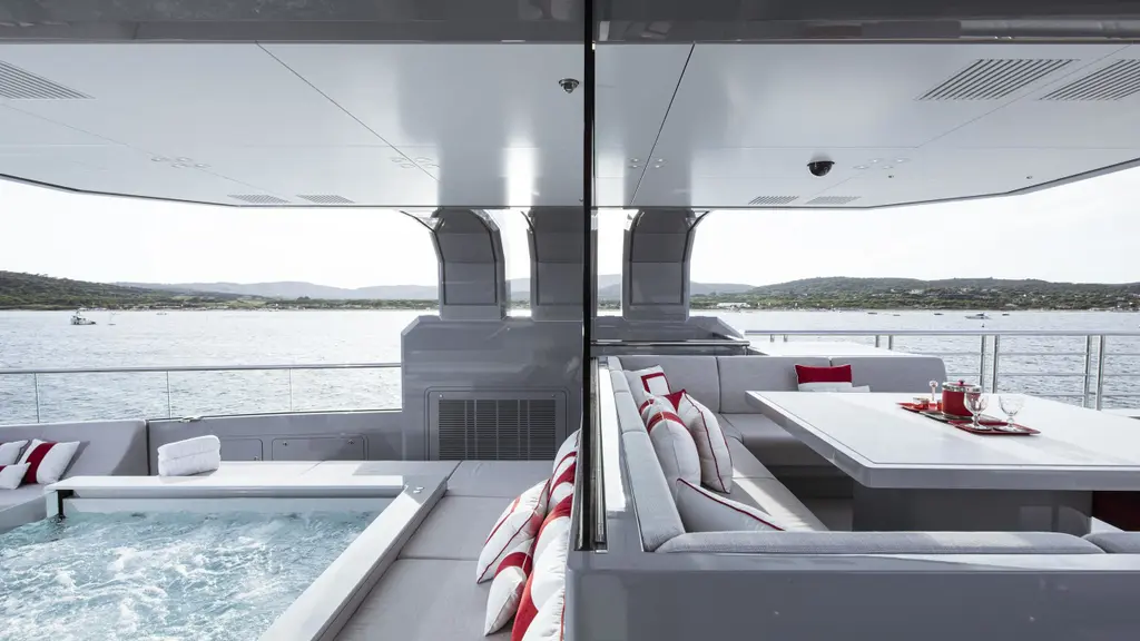 A glass-fronted spa pool on the sundeck under a hardtop, beside a lounge with red and white cushions