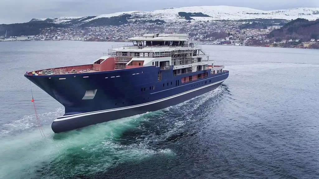 The dark blue hull of the yacht at speed off a snowy Norwegian town, with scaffolding still on the top deck