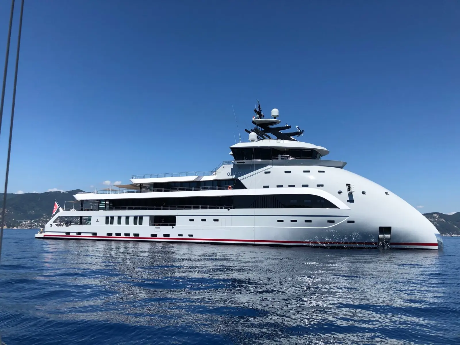 Olivia O at anchor in calm blue water, seen from the starboard side: the rounded inverted bow on the right, the glass bridge and mast above it, and the long white superstructure running aft