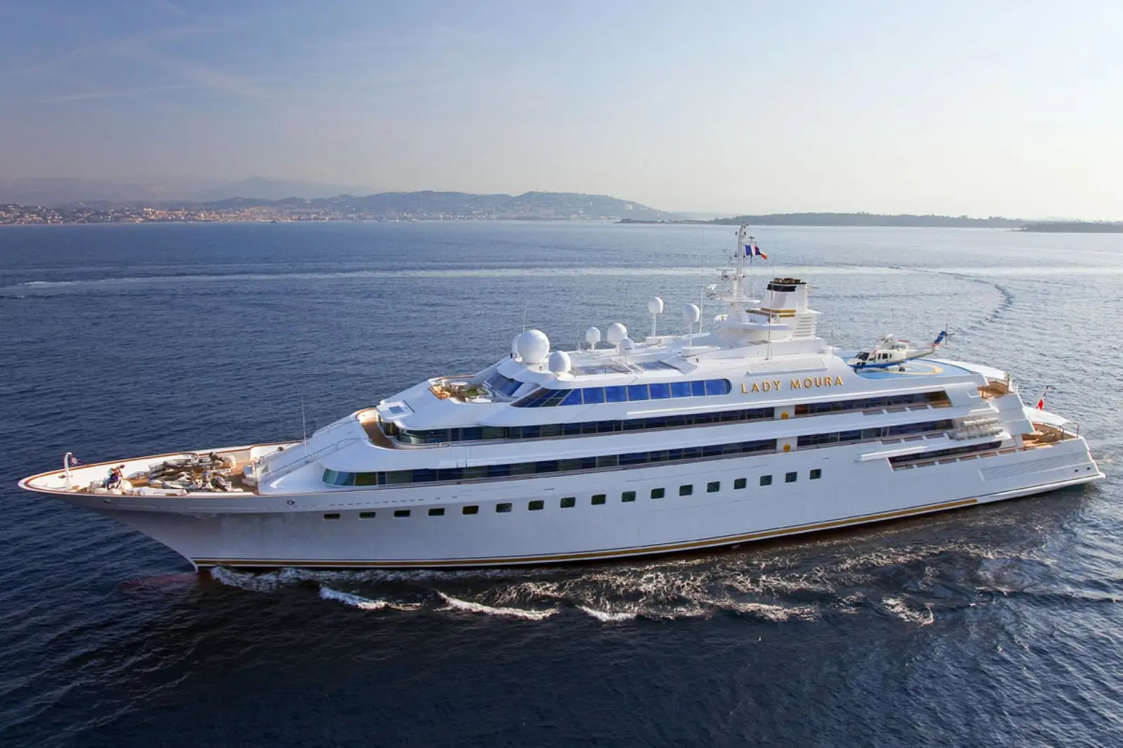Lady Moura under way, three-quarter view of the white hull and tiered upper decks, mountains on the horizon