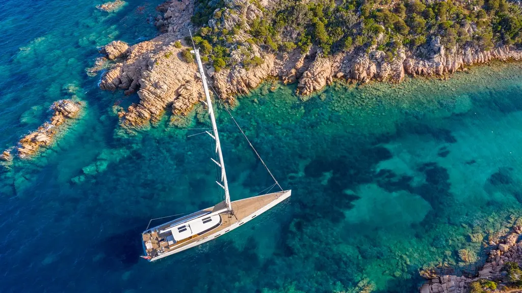 Canova at anchor in a turquoise bay below a rocky, wooded shore, seen from the air