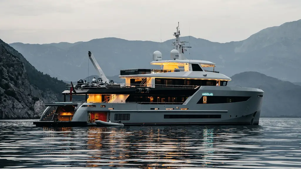 Heeus at anchor at dusk with her lights on, the crane raised over the boat deck, the beach club open at the stern and a tender alongside