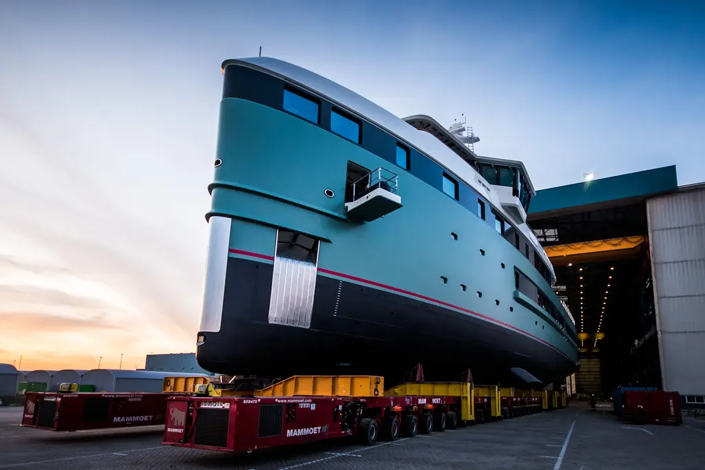 Anawa's turquoise hull on yellow and red self-propelled transporters outside a build hall in twilight, seen from the port bow with a steel bow fender and red stripe
