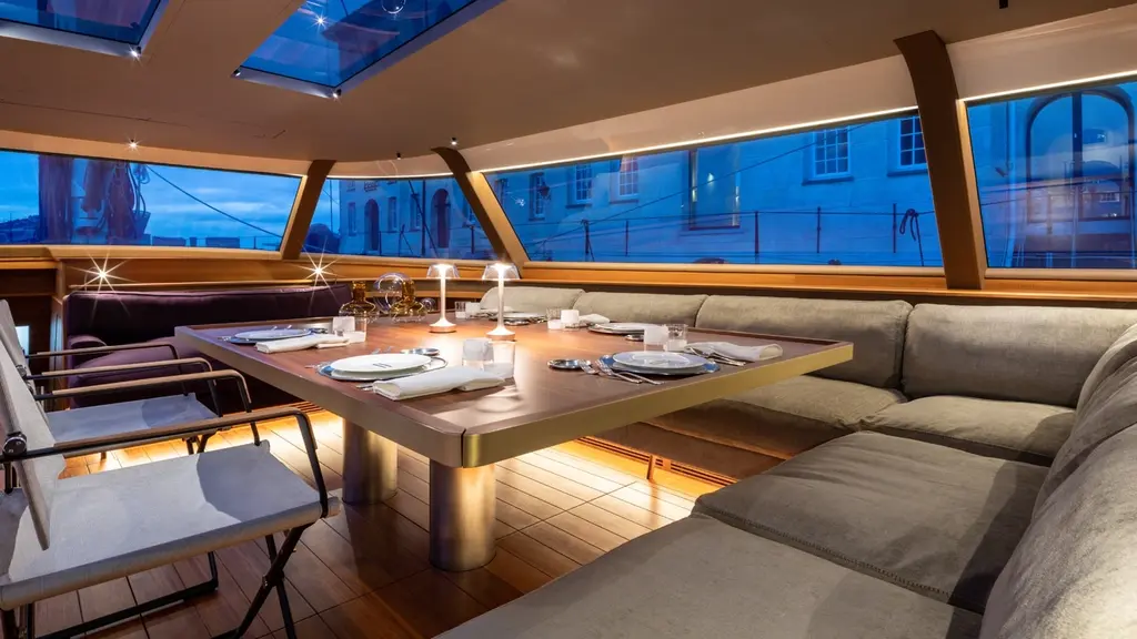 Dining table laid for dinner in the deck saloon, with grey sofas and windows on three sides