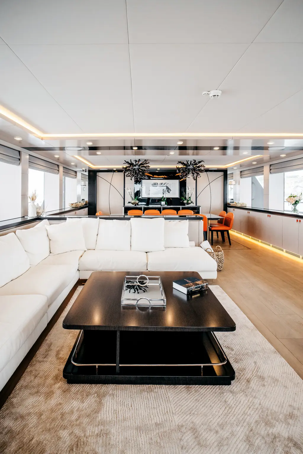 Main saloon with a white L-shaped sofa, a dark coffee table and the dining area with orange chairs beyond