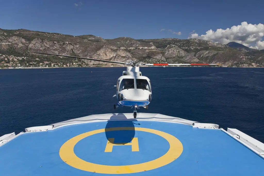 Helicopter hovering over Lady Moura's blue helideck, seen from the deck with the coast behind