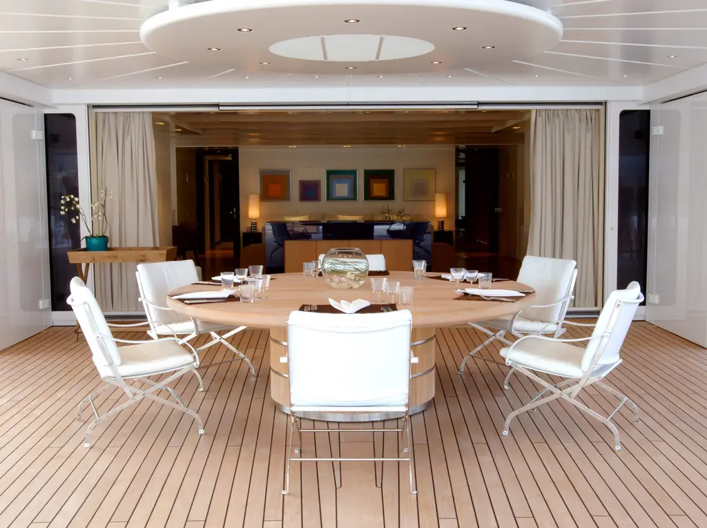 A round table with white chairs set for dinner on a covered teak deck in front of a lounge