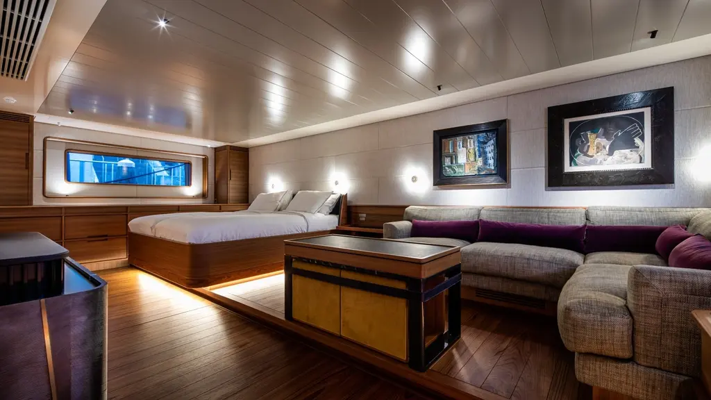 Large cabin with a double bed, a lounge sofa, a low table and framed art, lit by long hull windows