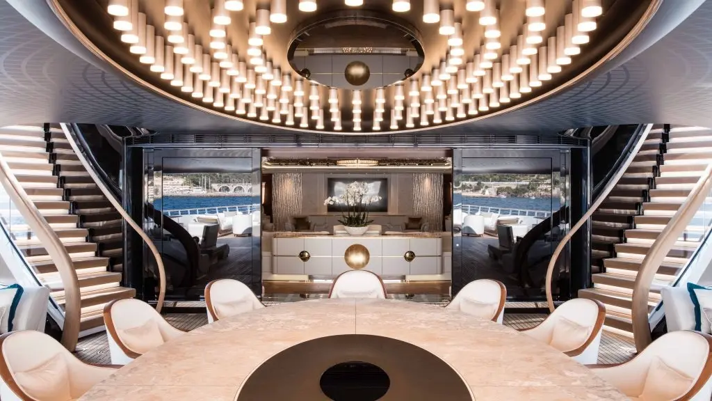 An oval dining table under a ring-shaped chandelier, with curving staircases on both sides