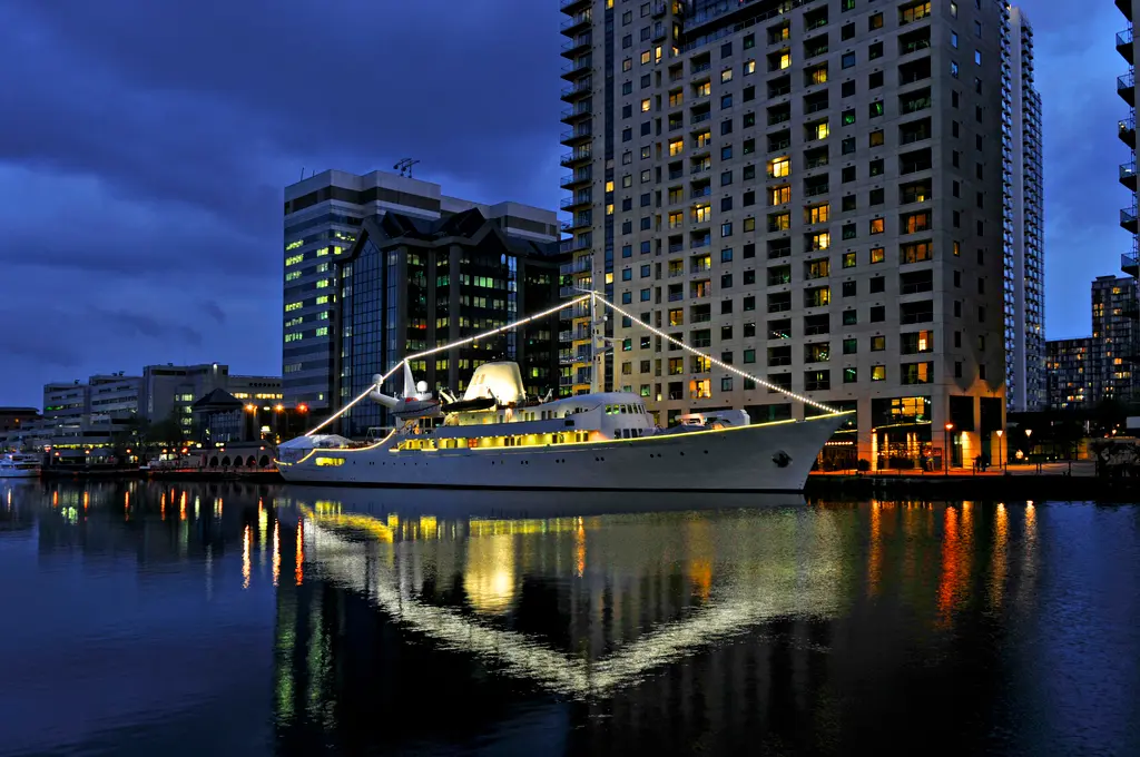 Christina O lit up at night alongside a city waterfront, reflected in the water