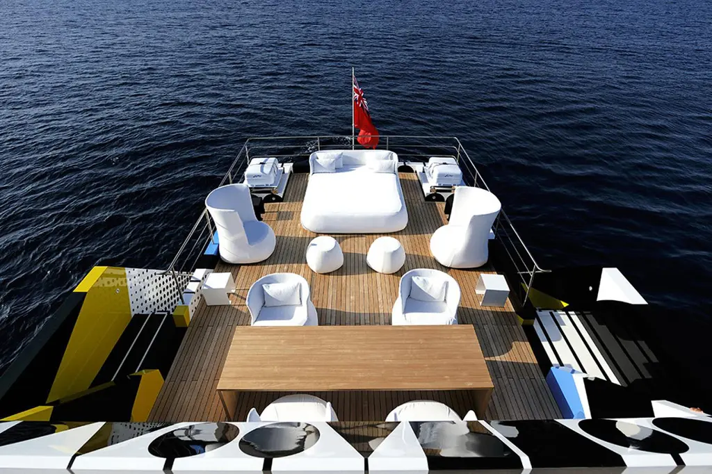 Open teak deck aft seen from above, with white armchairs, a large daybed and a long wooden table, the camouflaged hull at the edges