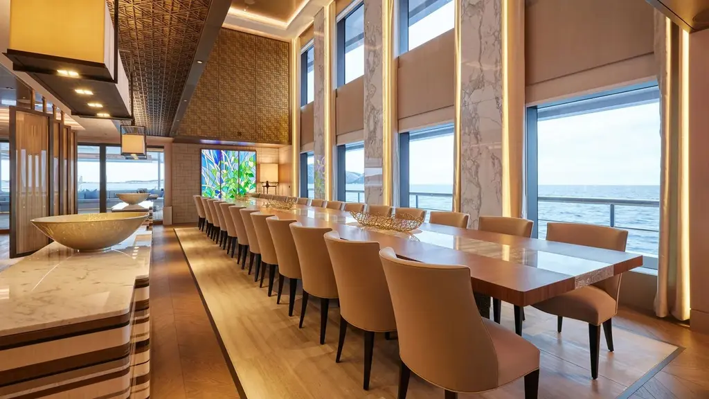 Long dining table with beige chairs beside tall windows to the sea