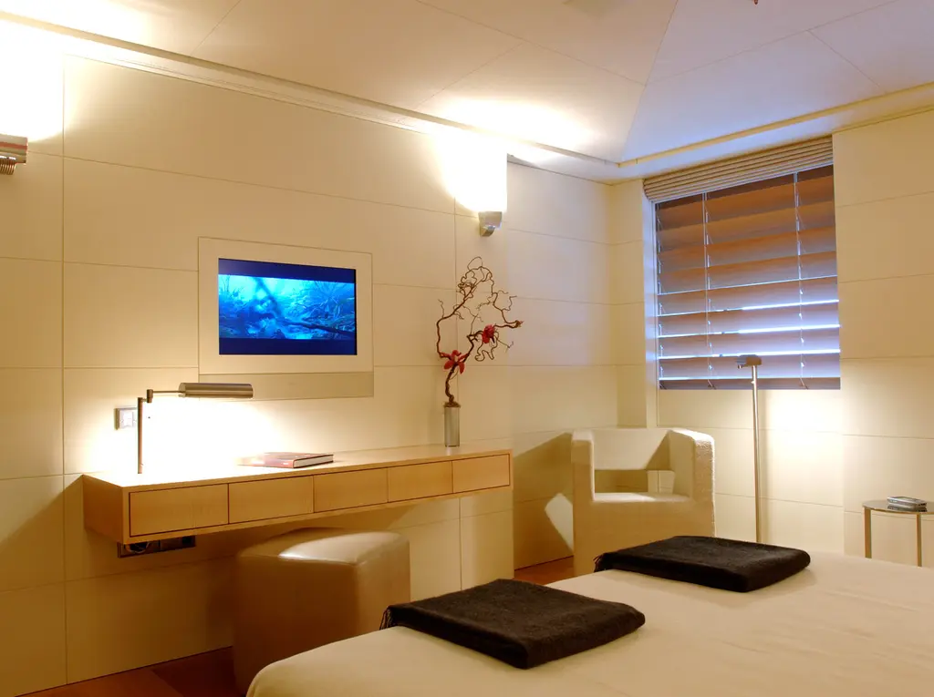 A guest cabin with a desk, a wall screen, blinds over the windows and a pale fabric bed