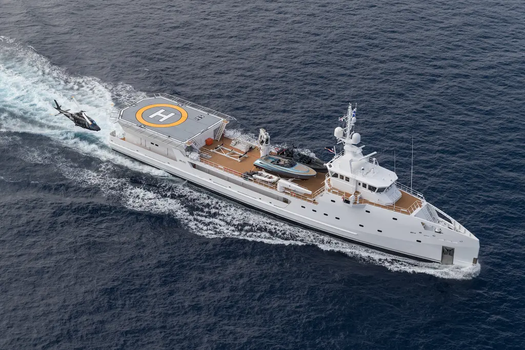 Aerial view of Game Changer at sea: the orange-marked H helideck aft, a teak working deck with a black tender amidships, and a helicopter flying alongside