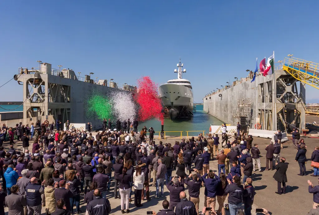 Crowd of shipyard workers at the launch ceremony, coloured smoke in green, white and red rising in front of the yacht's bow in the dock