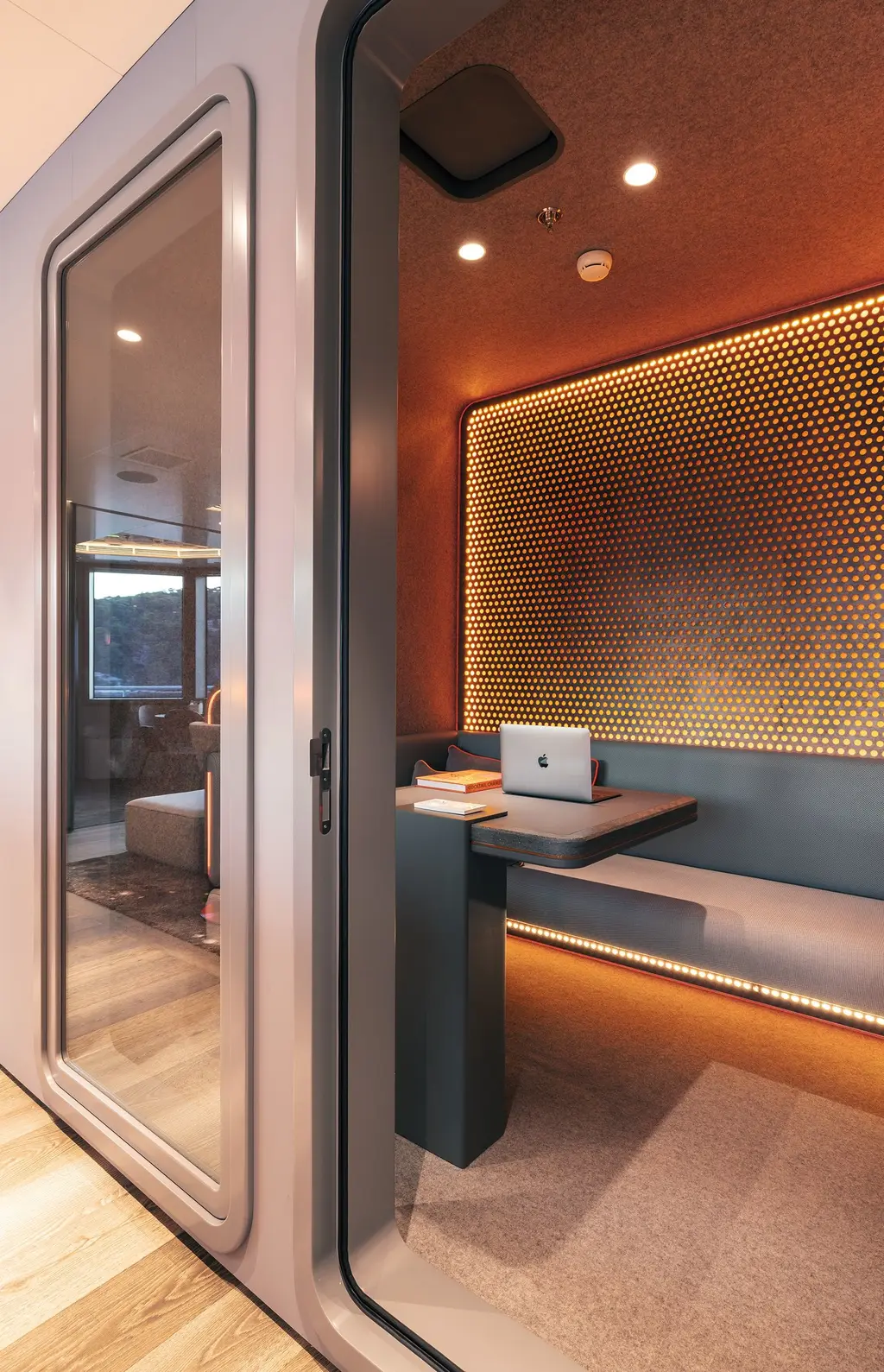 Inside a small soundproofed booth with orange walls, perforated lit panels, a bench and a desk with a laptop, seen through a glass door