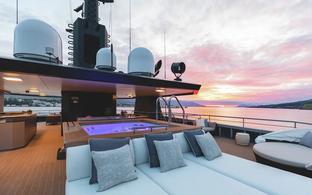 The top deck at dusk: a spa pool lit purple under a hardtop with satellite domes, sofas with blue cushions and a pink sky over a fjord
