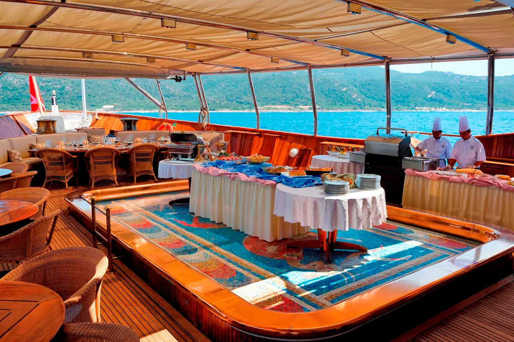 Aft deck under an awning set for a buffet, chefs at a grill, mosaic pool floor in front