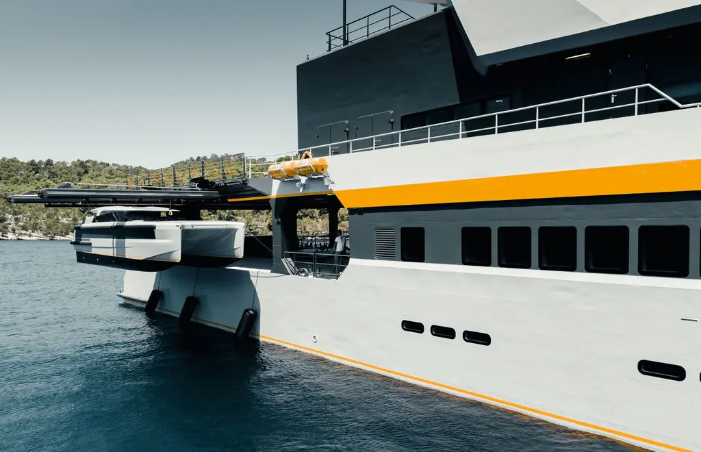 A white catamaran tender hanging from a beam crane over the water beside Nebula's starboard quarter, with the grey hull and orange stripe in the foreground