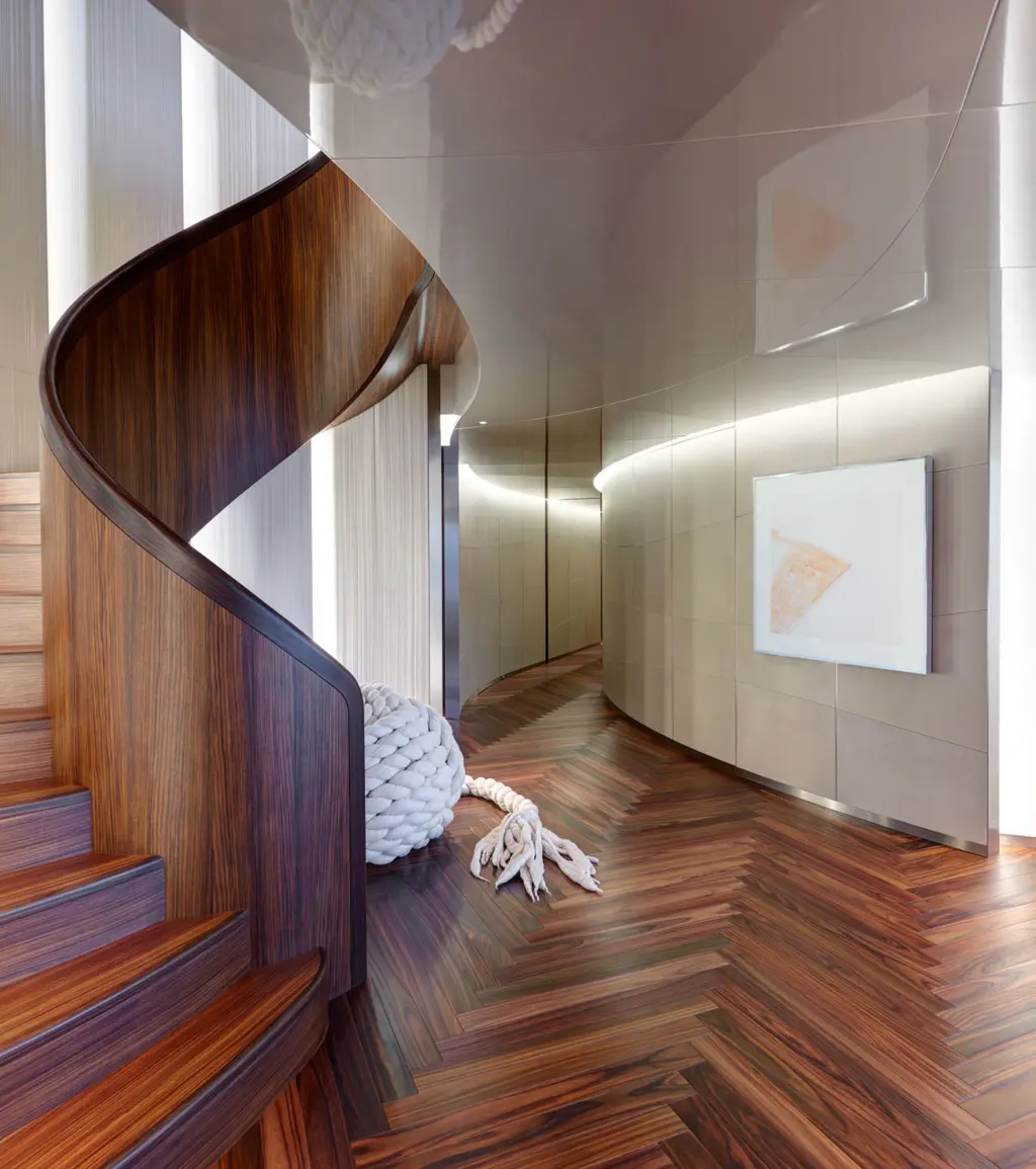 The curved rosewood staircase rising past a pale curved wall and a herringbone floor