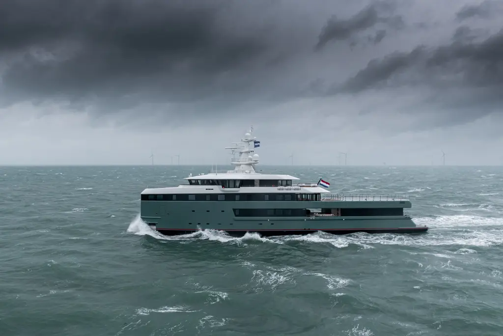 Anawa running through choppy sea under a dark storm sky, seen side-on from her port side, with wind turbines on the horizon and a Dutch flag flying aft of the superstructure