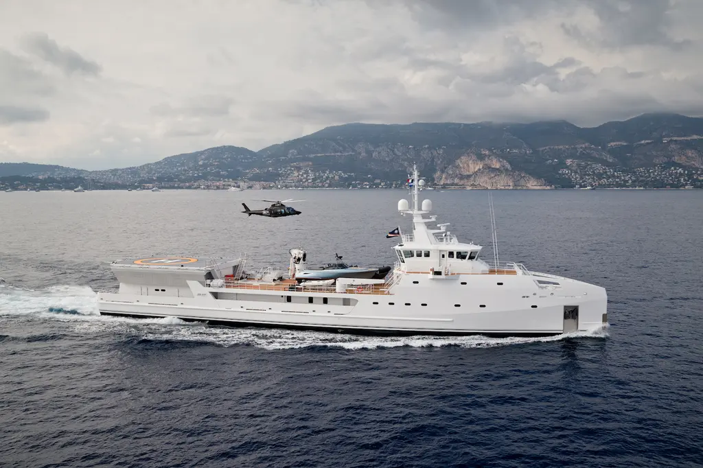 Game Changer under way along a mountainous coast in profile, with tenders on the working deck and a helicopter flying above
