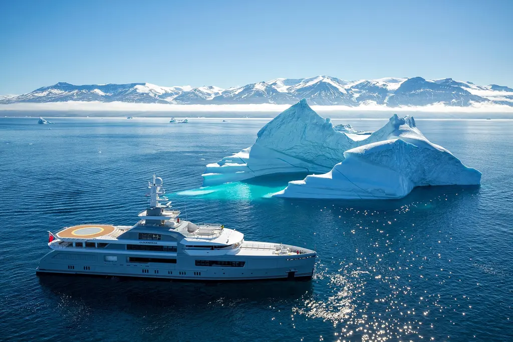 Aerial view of Cloudbreak in dark blue water near two large icebergs, with the orange-marked helideck visible on her upper deck and an icy coastline behind