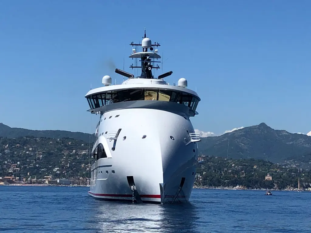 Olivia O seen head-on at anchor: the bulbous inverted bow rising to the bridge, with wing stations either side and a wooded coastline behind