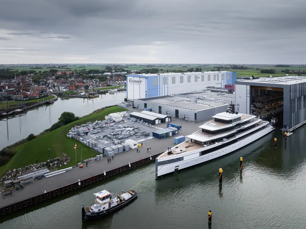 Aerial view of Ulysses alongside the Feadship yard quay, with a tug in the foreground and the build halls behind