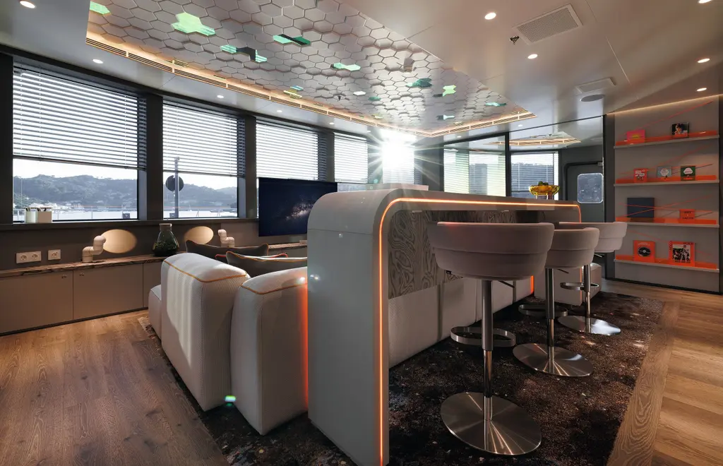 Nebula's crew lounge with a curved white bar and stools, a sofa, orange-lit shelving and a ceiling of hexagonal felt panels