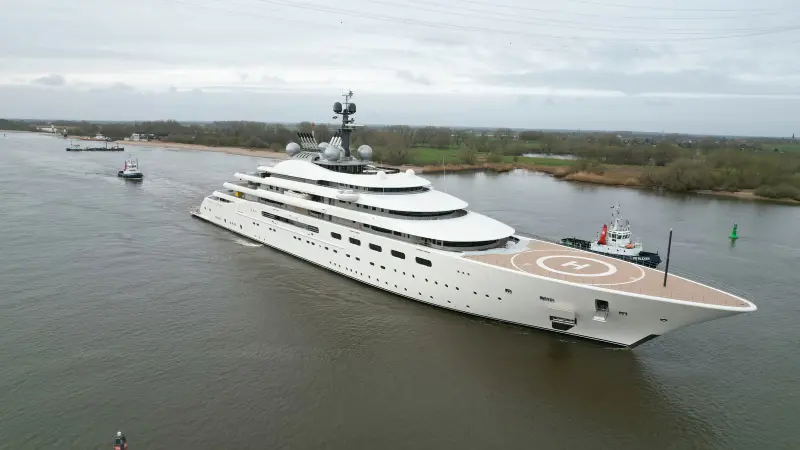 Blue underway on a wide river under a grey sky, helipad on the foredeck, a tug alongside