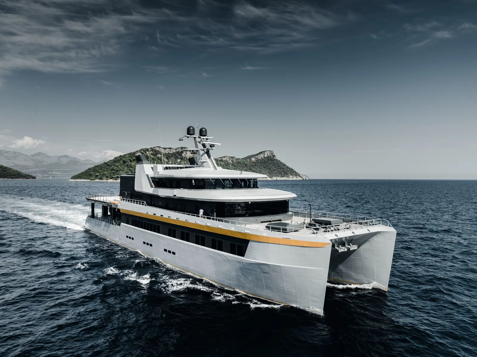 Nebula under way in open water off a green island, seen from ahead on her starboard bow: a light grey catamaran with an orange stripe, a dark superstructure and a mast with radar domes