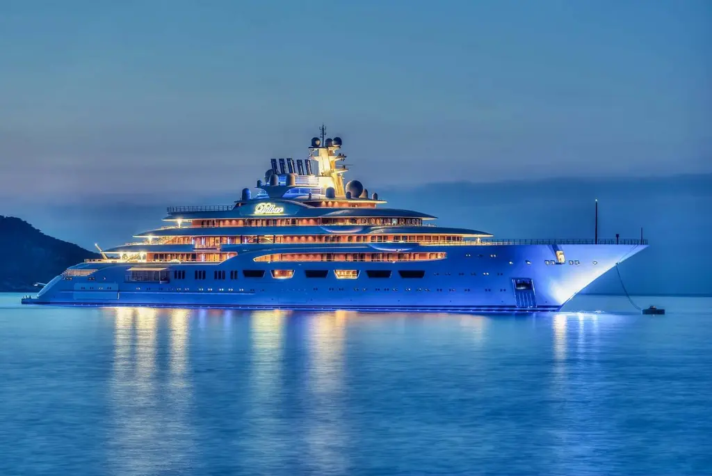 Dilbar at anchor at dusk with her deck lights on, reflected in calm water
