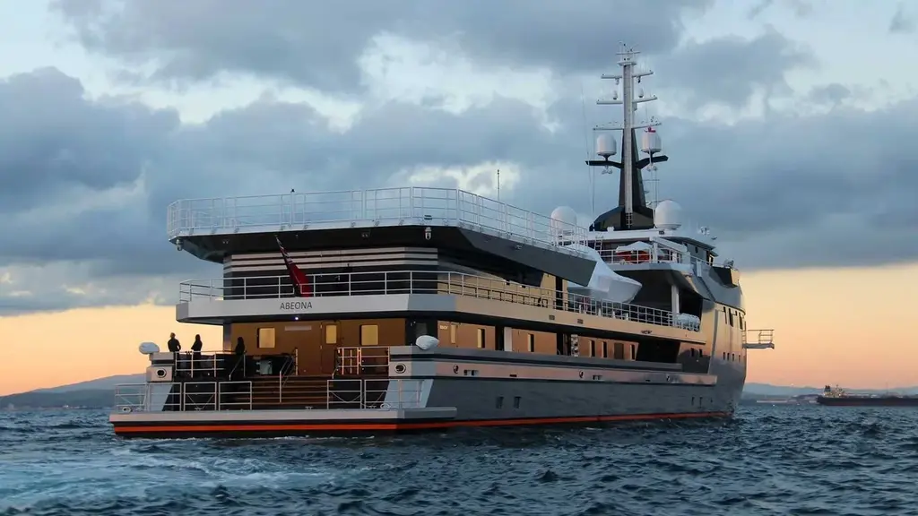 The stern of Abeona with her name on the superstructure, the railed helideck on top, tenders under covers and crew on the aft deck at dusk
