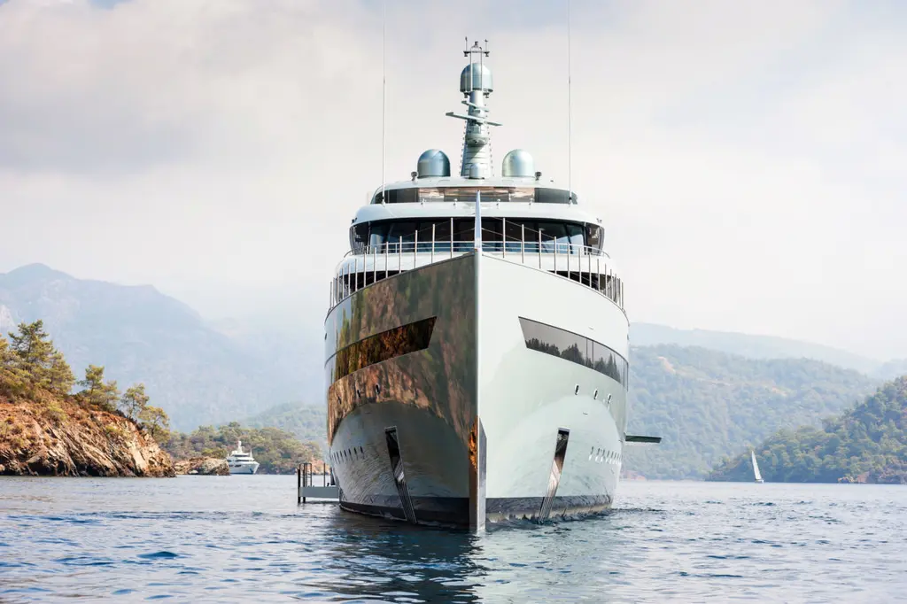 Savannah at anchor seen head-on, the metallic hull reflecting the water, wooded hills behind