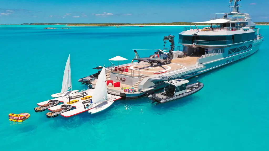 Bold at anchor in shallow turquoise water with a helicopter on the aft deck, sailing dinghies, jet skis and tenders around a floating dock at the stern