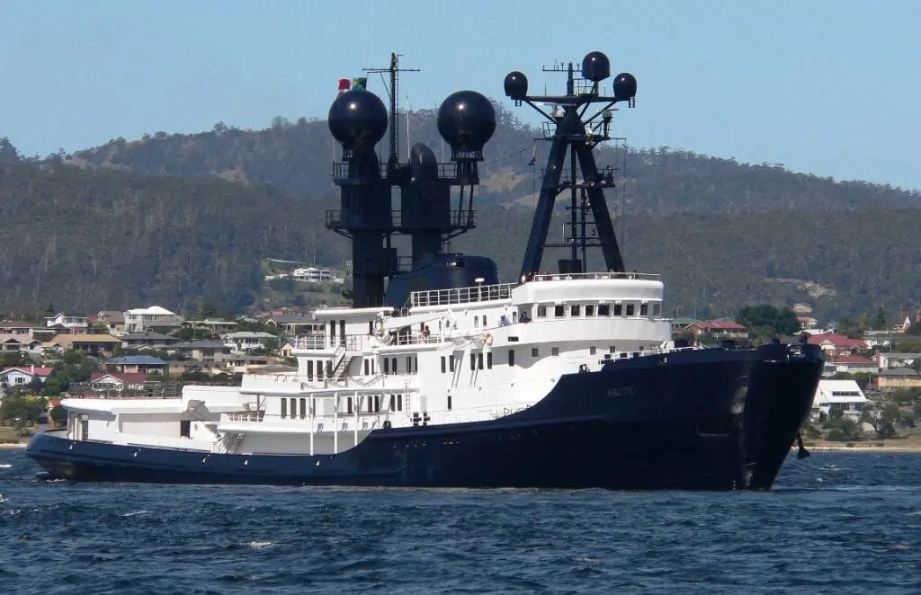 Arctic P at anchor off a wooded hillside with houses: the high black tug bow on the right, white superstructure and two tall black masts with radar domes
