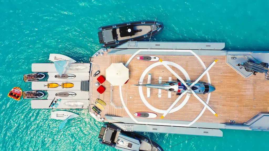 Overhead view of the aft deck at anchor in turquoise water, with a helicopter parked on the marked landing circle, tenders alongside and a floating dock with jet skis