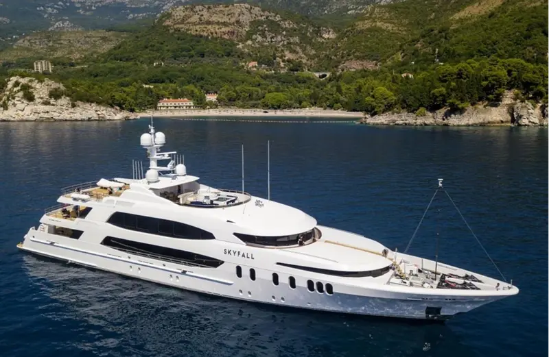 Skyfall at anchor in a bay, white hull and superstructure seen from above the starboard bow, wooded hills behind